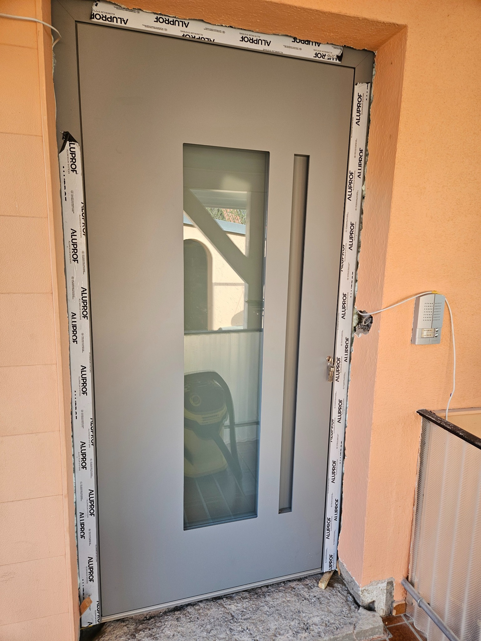 Szare drzwi zewnętrzne z szybą, widoczne ślady montażu i pianka uszczelniająca wokół ościeżnicy, na ścianie domofon.