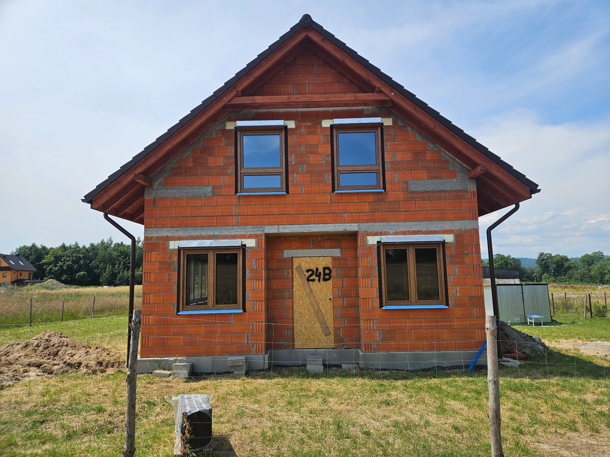 Niedokończony budynek z czerwonej cegły z częściowo zamontowanymi oknami z brązowymi ramami, zabezpieczonymi niebieską folią, i deskami zamiast drzwi wejściowych z napisem 24B, na tle trawnika i drzew