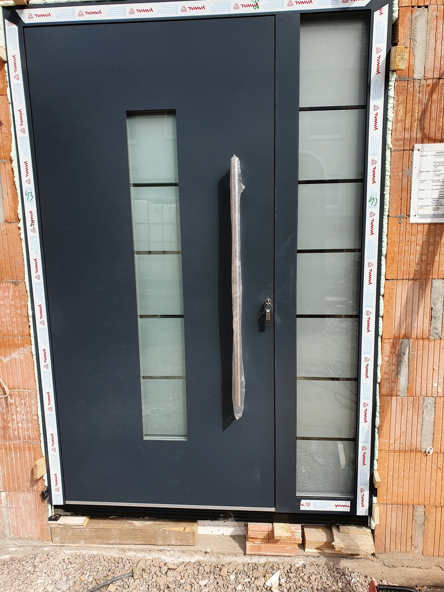 Nowoczesne, grafitowe drzwi zewnętrzne z przeszkleniami, zamontowane w surowym murze z cegły, widoczne zabezpieczenie klamki folią ochronną.