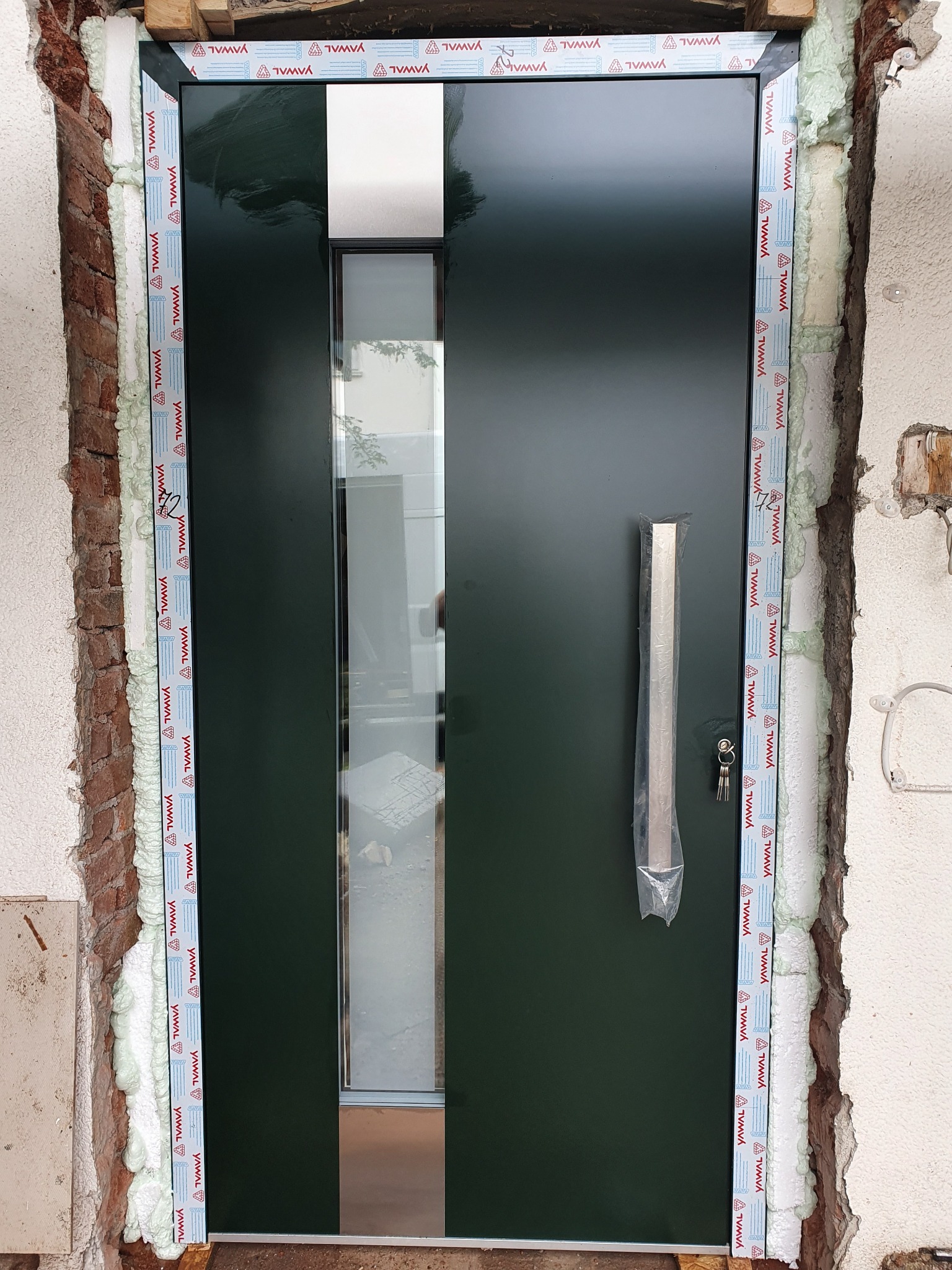 Ciemnozielone drzwi wejściowe z długim, pionowym przeszkleniem i srebrnym uchwytem w folii ochronnej, osadzone w surowym otworze ściennym z widoczną pianką montażową i elementami muru.