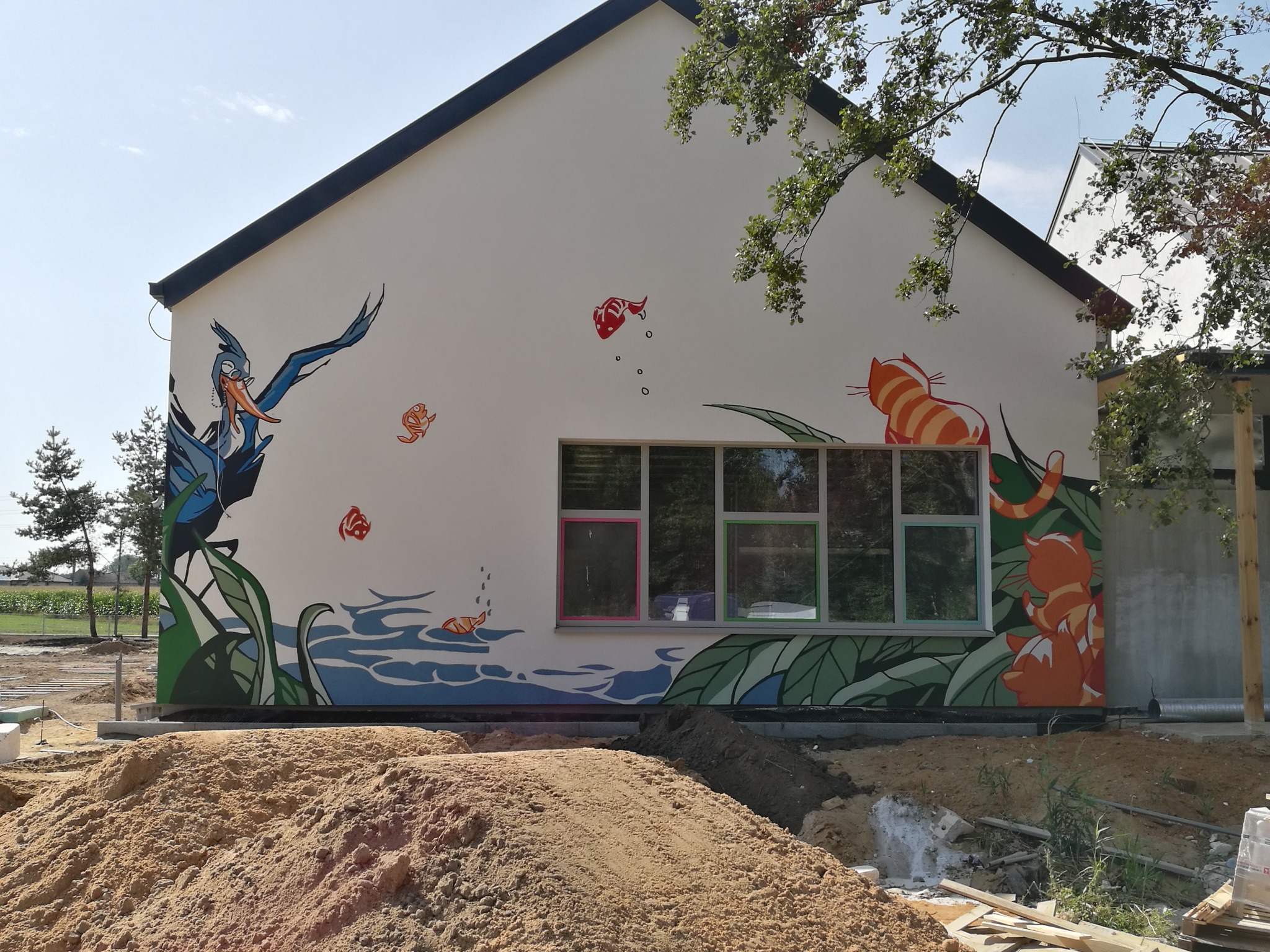 Ściana budynku z kolorowym muralem przedstawiającym ptaka, ryby i koty, okna z ramami w kolorach różowym i zielonym, widoczny plac budowy.