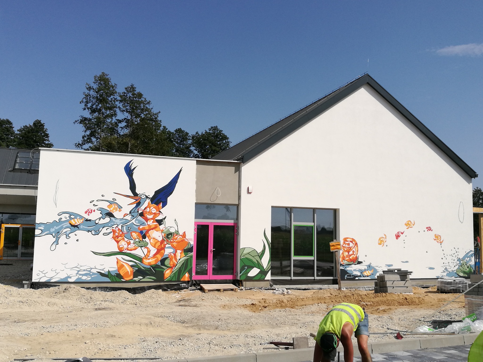 Nowoczesny budynek z białymi ścianami ozdobiony kolorowymi muralami z motywami zwierzęcymi i roślinnymi, różowe drzwi wejściowe, widoczne prace wykończeniowe na zewnątrz.