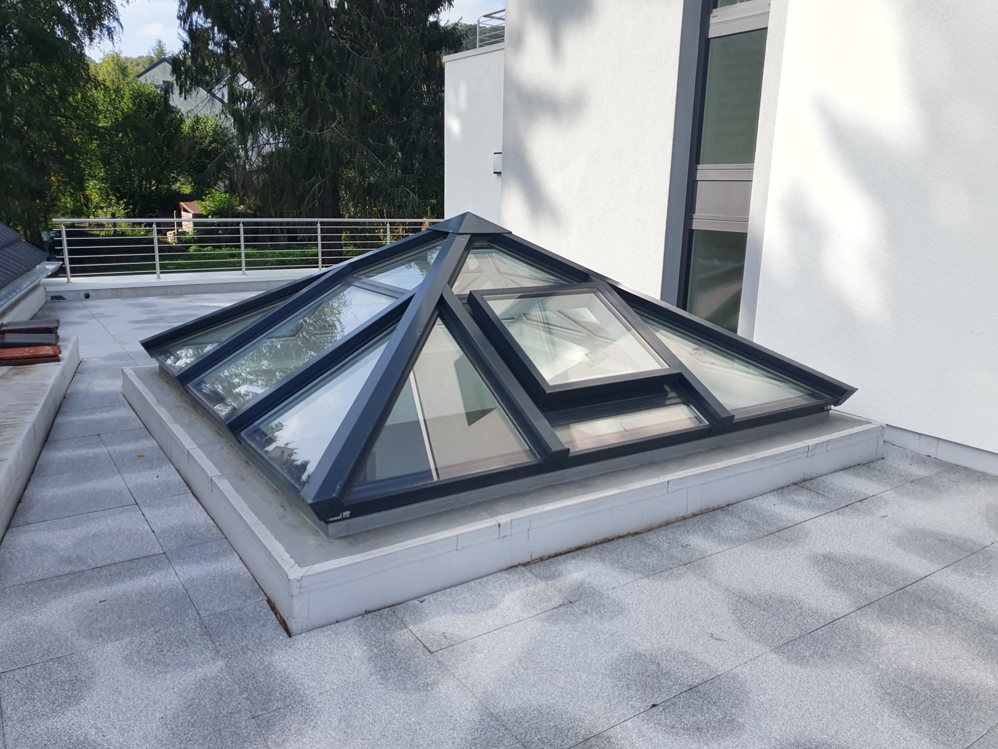 Piramidalny świetlik dachowy z czarną ramą na płaskim dachu z szarych płyt, widok z góry, w tle biała ściana budynku i zieleń drzew.