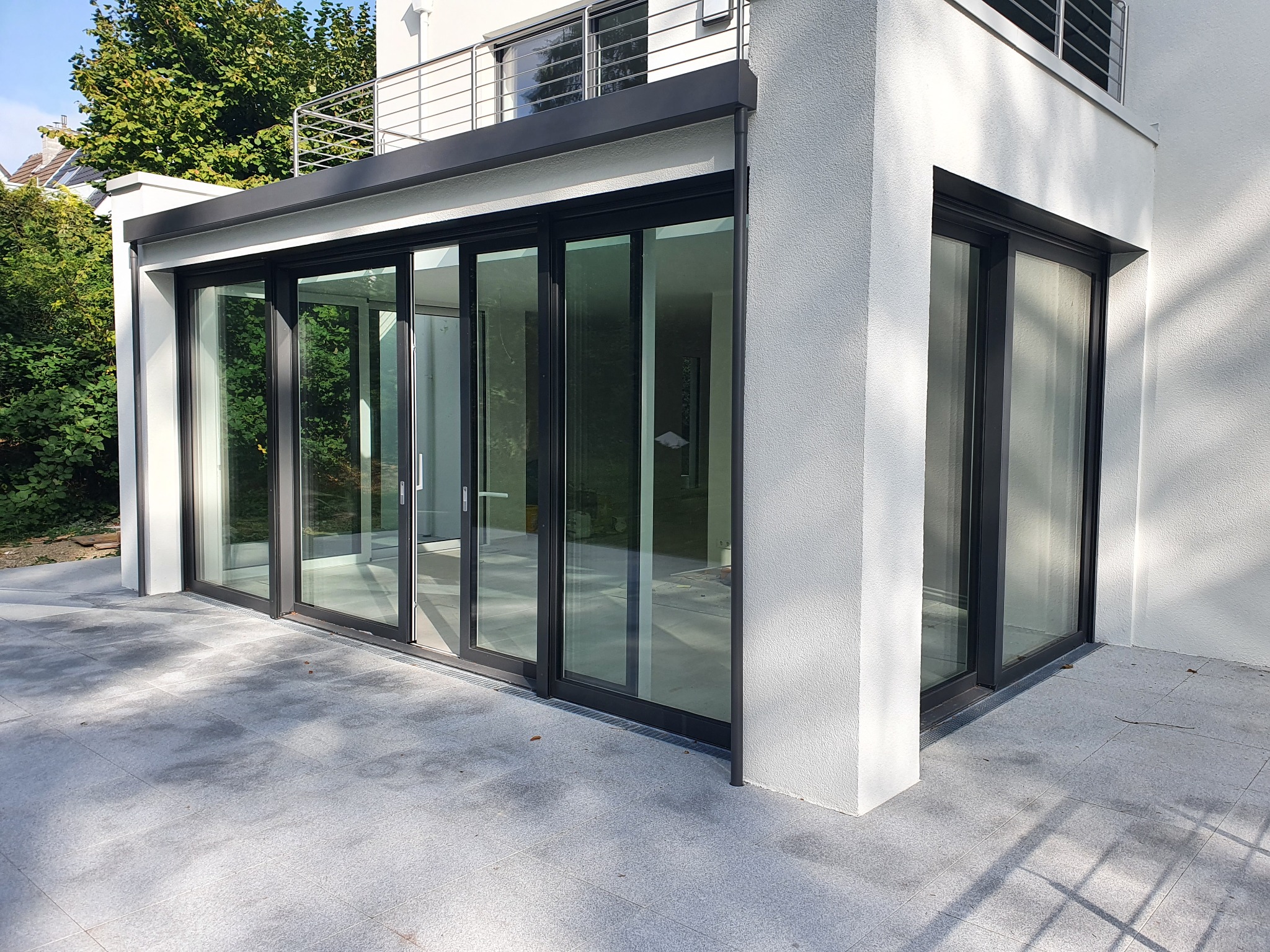 Nowoczesny przeszklony taras z przesuwnymi drzwiami, czarne ramy, szara posadzka z płytek, widok z zewnątrz, balkon na górze.