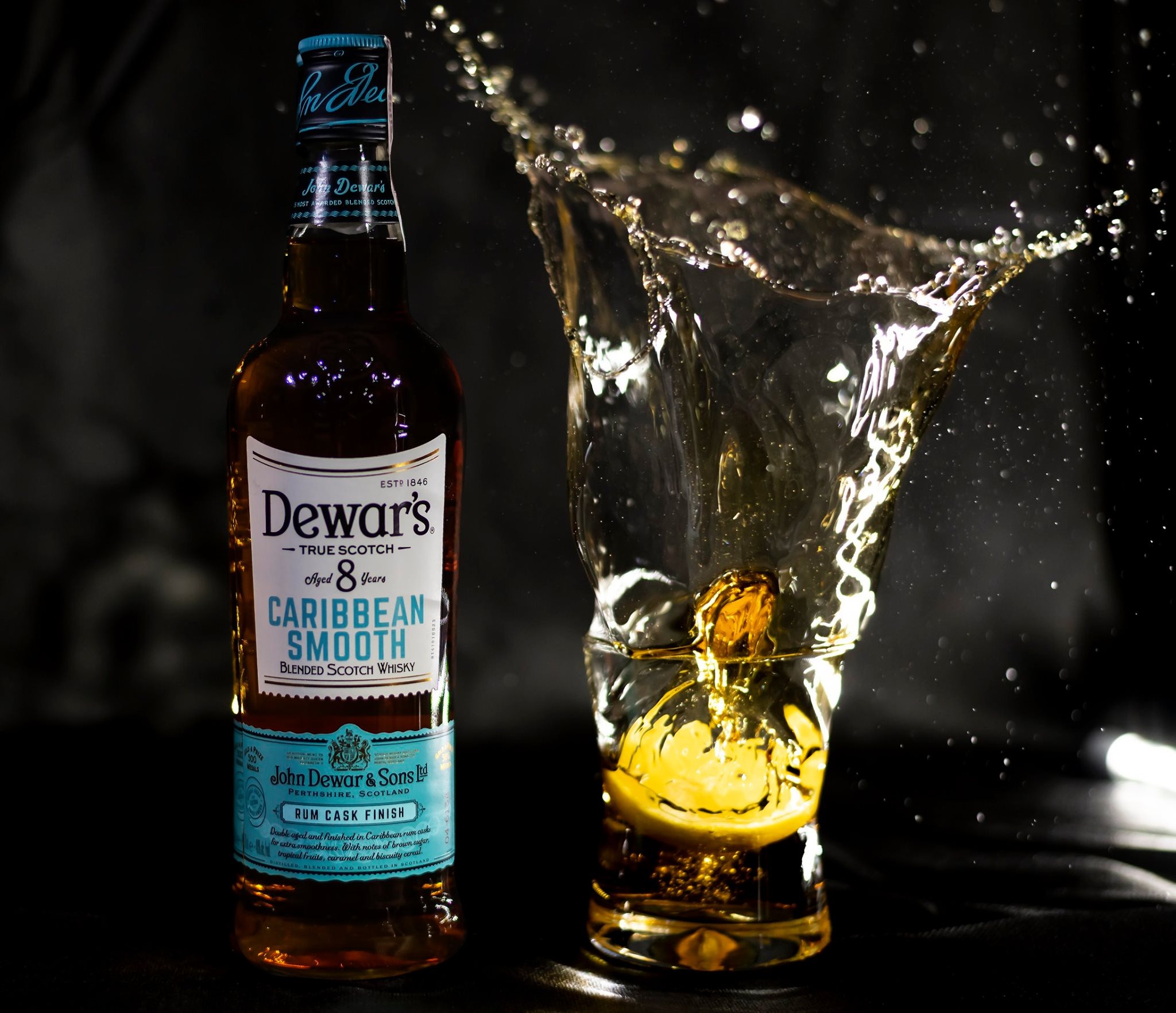 Butelka whisky Dewar's Caribbean Smooth obok szklanki, do której wpadła kostka lodu, powodując rozprysk płynu na czarnym tle.