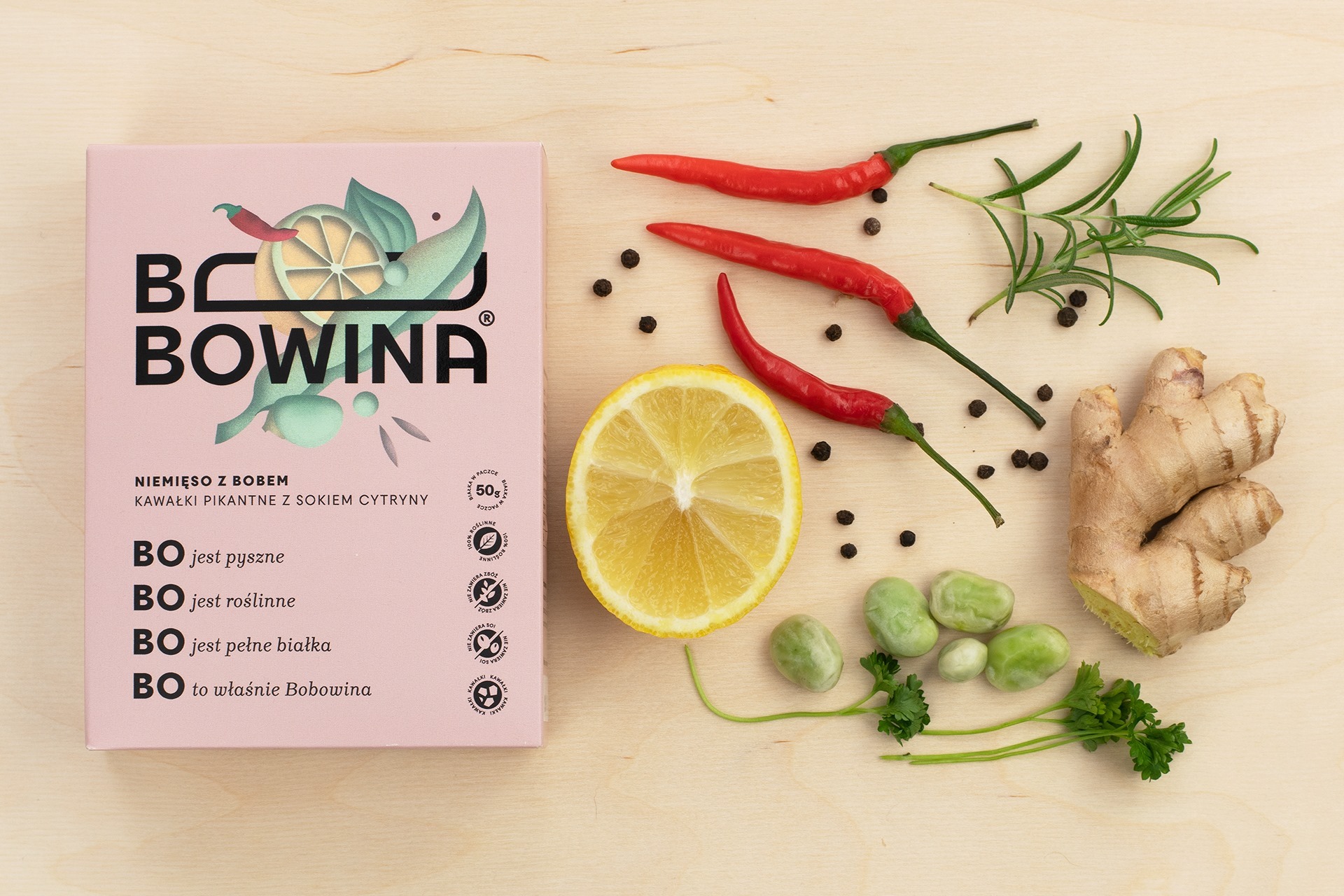 Różowe opakowanie Bobowiny z napisem 'Niemi ęso z bobem, kawałki pikantne z sokiem cytryny', obok połówka cytryny, trzy czerwone papryczki chili, ziarna czarnego pieprzu, gałązka rozmarynu, korzeń...