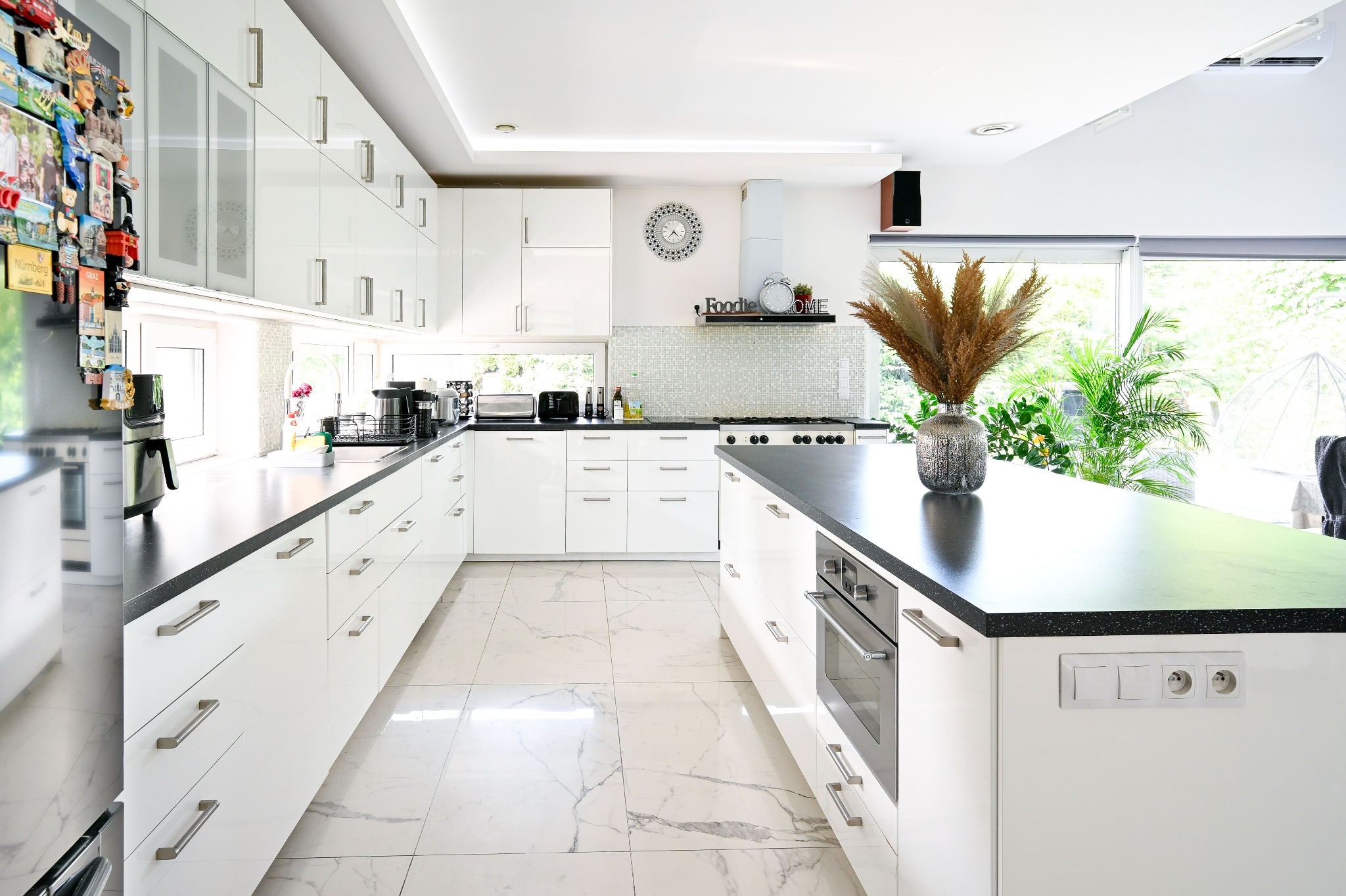 Jasna, nowoczesna kuchnia z białymi szafkami, czarnymi blatami i marmurową podłogą; wysoka wyspa kuchenna z piekarnikiem, dekoracyjna trawa pampasowa w srebrnym wazonie, okno z widokiem na zieleń.