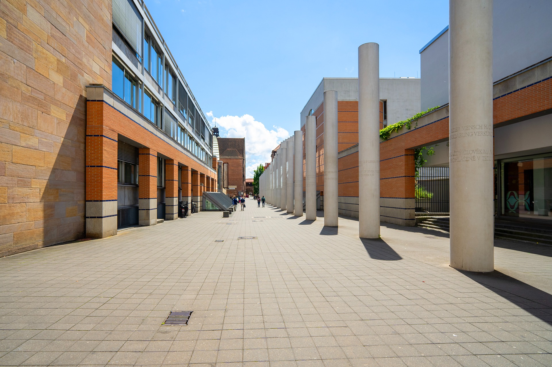 Zewnętrzny widok na architekturę miejską z rzędem kolumn z inskrypcjami, budynkami z cegły i piaskowca oraz pieszymi w oddali.