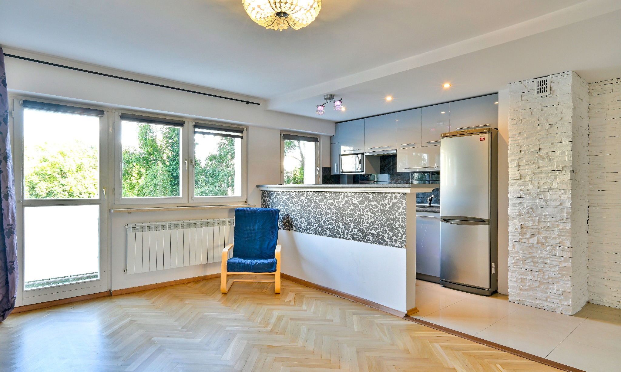Jasne, przestronne wnętrze mieszkania z parkietem w jodełkę, niebieskim fotelem i widokiem na zieleń za oknem; kuchnia z szarymi szafkami i lodówką ze stali nierdzewnej.