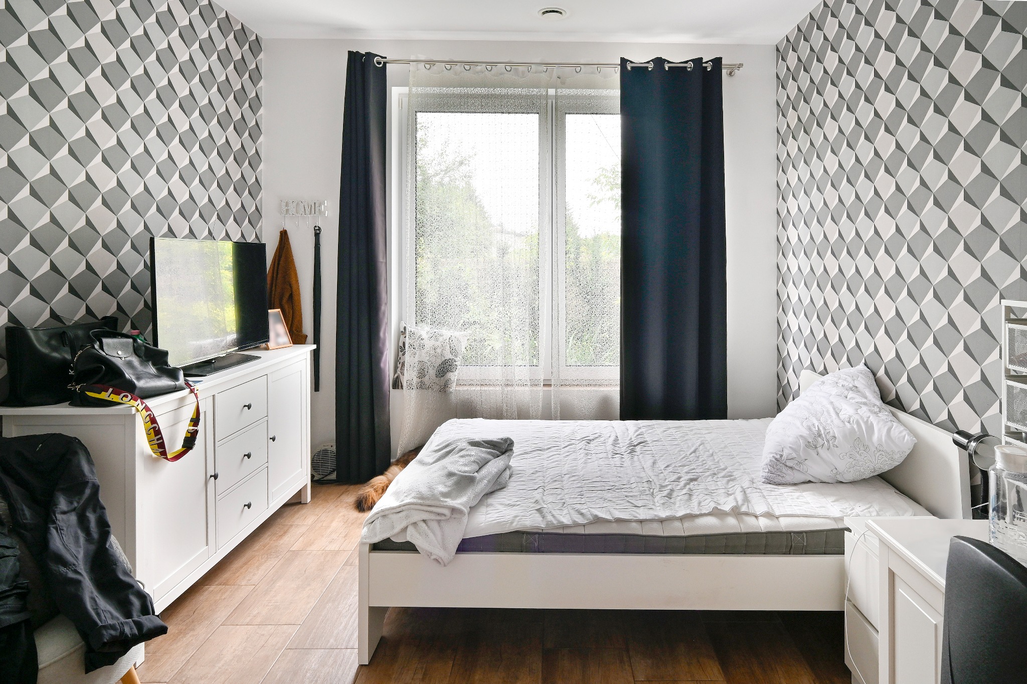 Pokój z łóżkiem, białą komodą z czarnymi uchwytami, telewizorem, wzorzystą tapetą w odcieniach szarości i oknem z ciemnymi zasłonami.