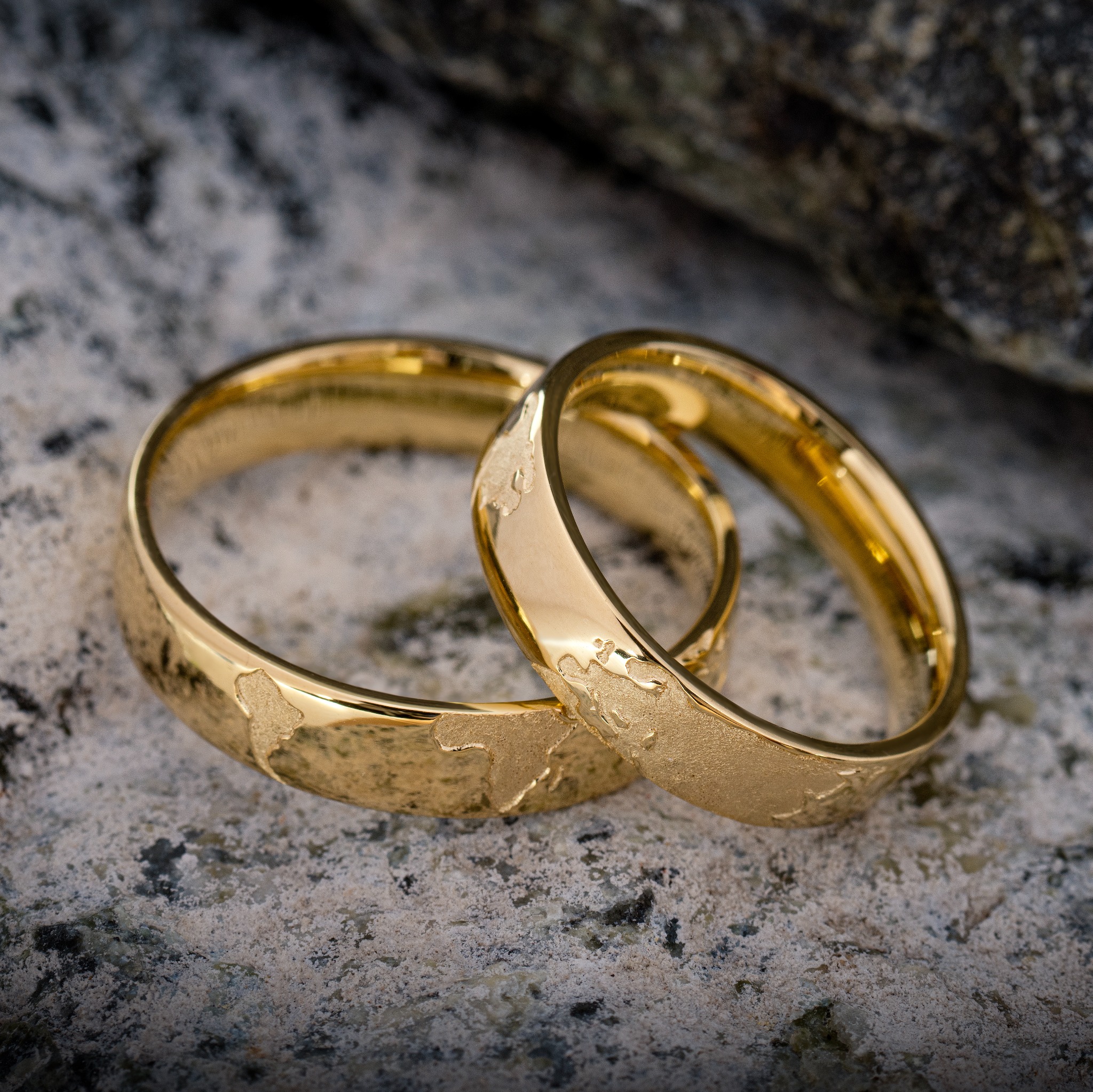 Dwa złote obrączki z wytłoczonymi konturami mapy świata leżące na szorstkiej, kamiennej powierzchni.