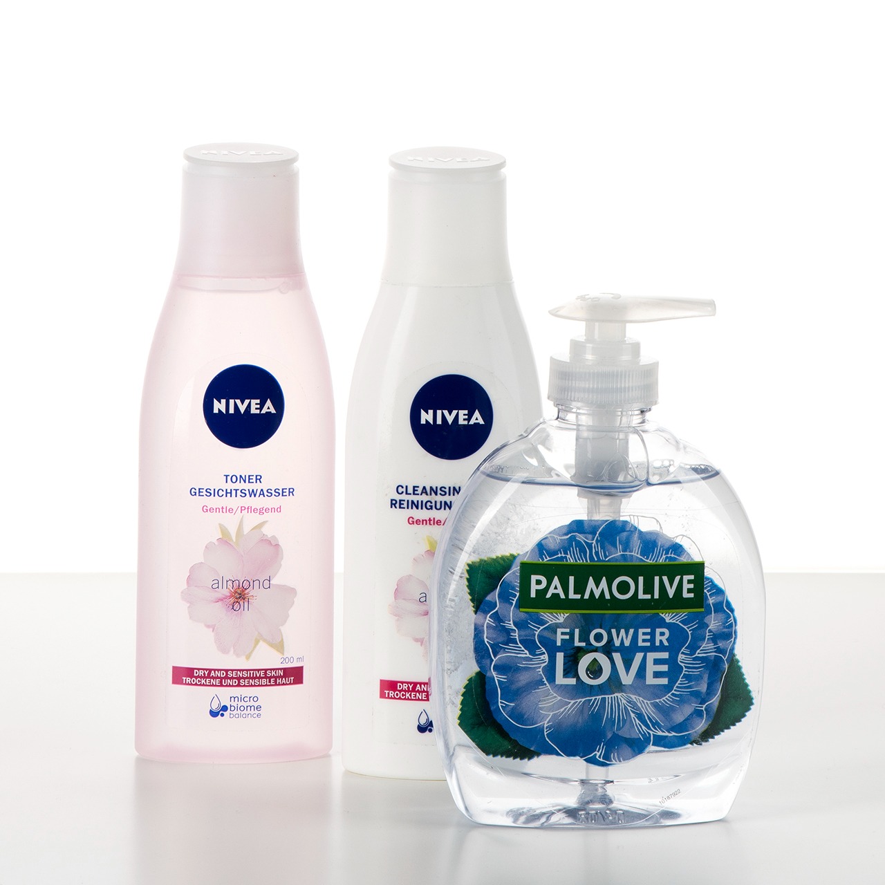 Trzy butelki kosmetyków: różowy tonik Nivea z olejkiem migdałowym, biały płyn do demakijażu Nivea oraz mydło w płynie Palmolive Flower Love w przezroczystej butelce z niebieskim kwiatem.