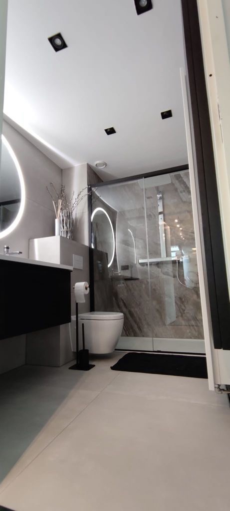 Nowoczesna łazienka z szarymi płytkami, kabiną prysznicową ze szklaną ścianą, okrągłym lustrem z podświetleniem LED i wiszącą toaletą z czarnym uchwytem na papier toaletowy.