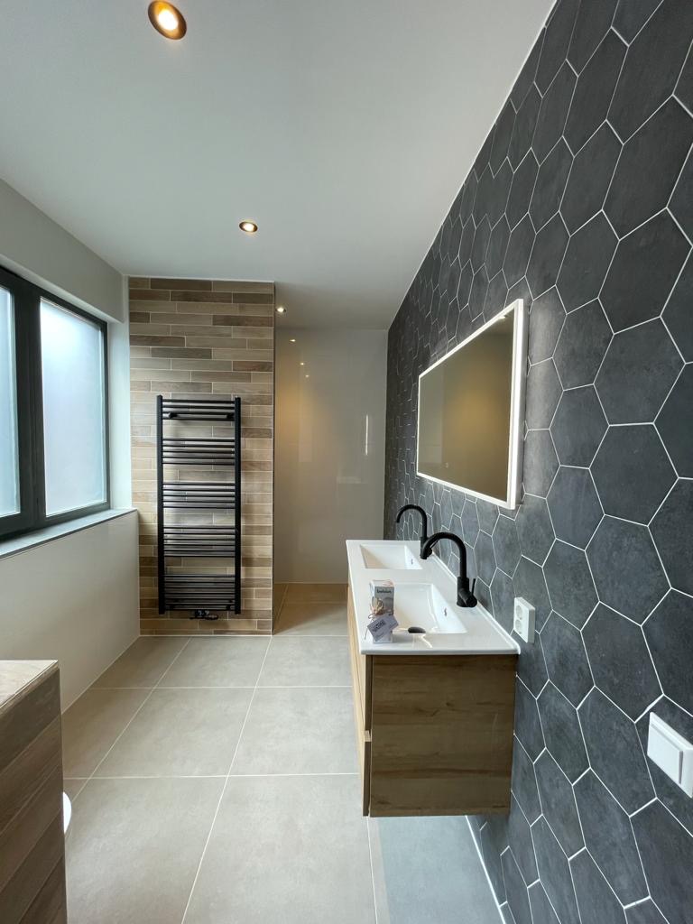 Nowoczesna łazienka z heksagonalnymi płytkami grafitowymi na ścianie, drewnianą szafką pod umywalką z dwiema białymi umywalkami i czarnymi bateriami, oraz grzejnikiem drabinkowym na ścianie...
