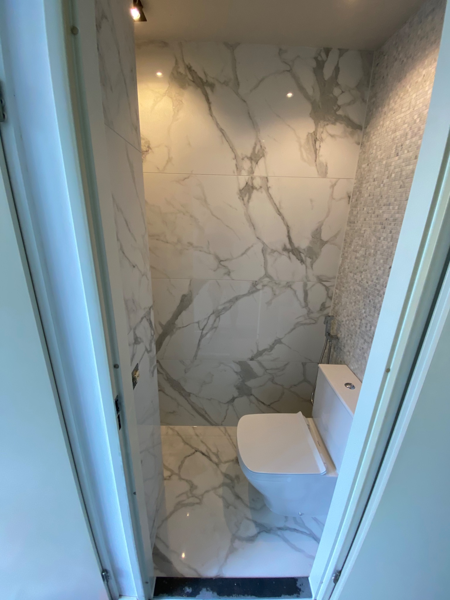 Wąska łazienka z toaletą, wykończona płytkami imitującymi marmur na podłodze i ścianach oraz mozaiką na jednej ze ścian, widok zza uchylonych drzwi.