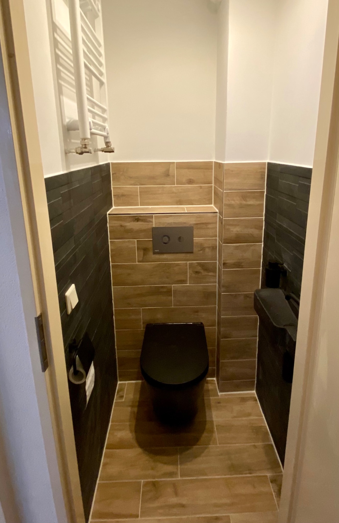 Wąska toaleta z czarną miską ustępową i drewnopodobnymi płytkami na podłodze i części ścian, kontrastującymi z ciemnymi, wąskimi płytkami na pozostałej części ścian. Grzejnik drabinkowy po lewej...