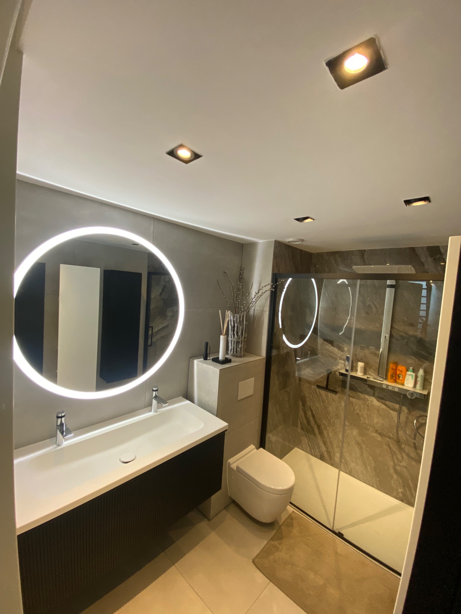 Nowoczesna łazienka z podświetlanym okrągłym lustrem nad podwójną umywalką, toaleta typu Geberit i kabina prysznicowa z imitacją kamienia na ścianach.