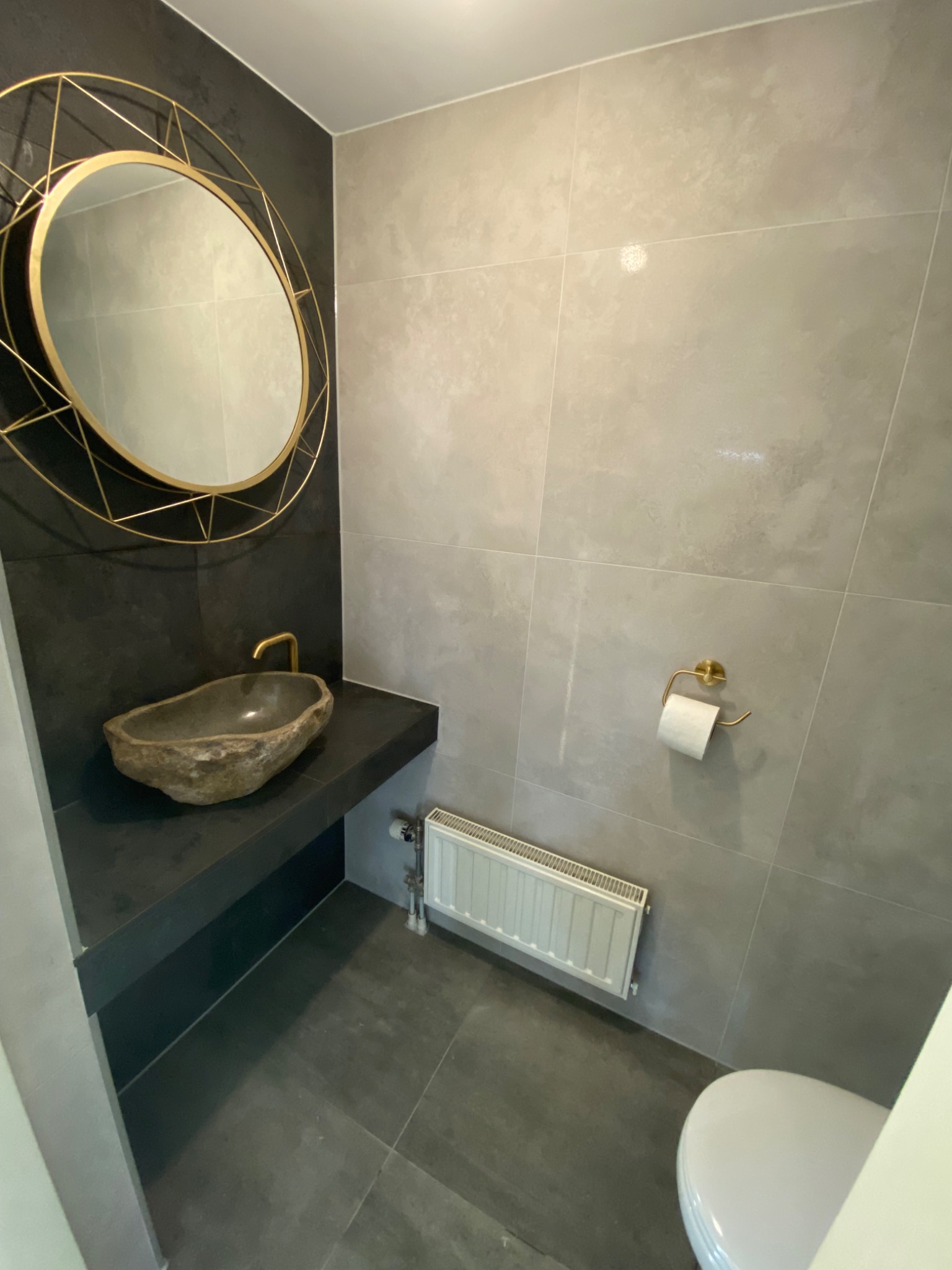 Nowoczesna łazienka z umywalką przypominającą kamień rzeczny, okrągłym lustrem w złotej ramie geometrycznej i szarą płytką na ścianach. Widoczny fragment toalety.