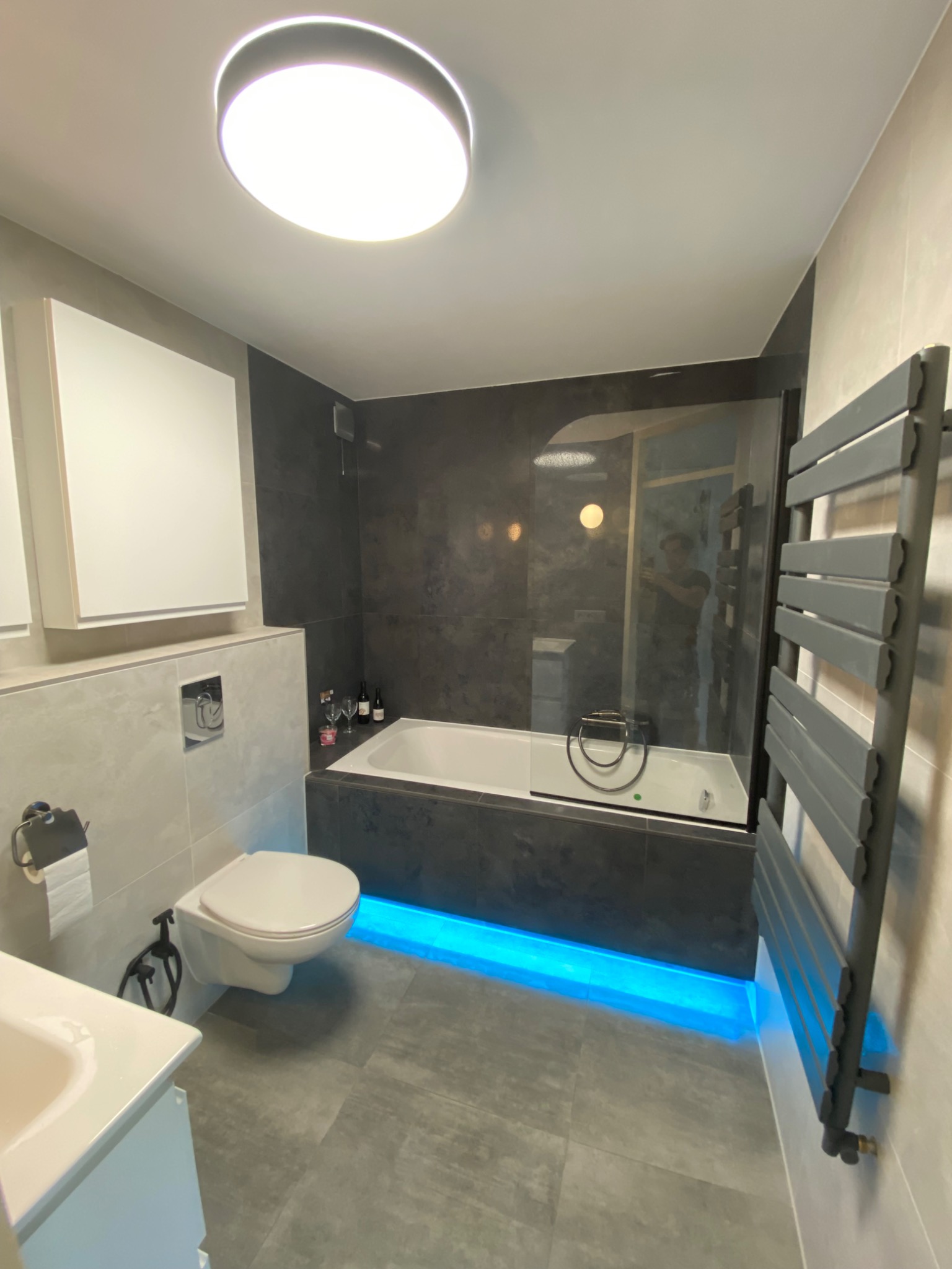 Nowoczesna łazienka z szarymi płytkami, podświetlaną na niebiesko wanną z parawanem, grzejnikiem drabinkowym i wiszącą toaletą z bidetką, uzupełniona o wino i kieliszki na obudowie wanny.