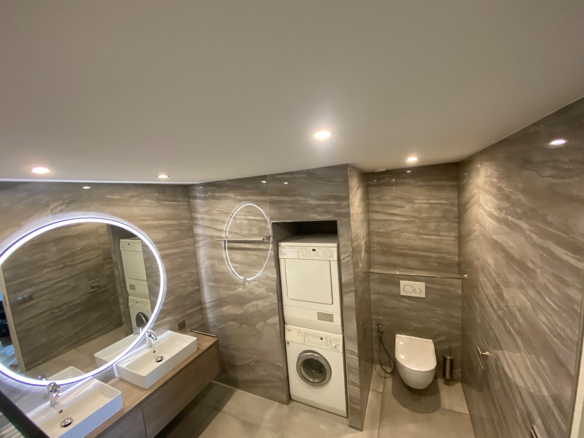 Nowoczesna łazienka z marmurowymi płytkami, okrągłym lustrem z podświetleniem LED, wiszącą toaletą oraz pralko-suszarką w zabudowie.