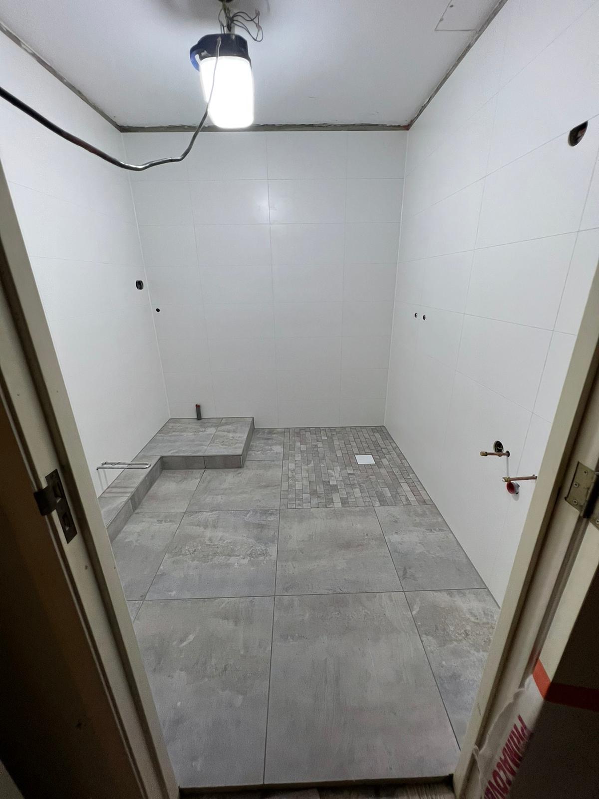 Łazienka w trakcie remontu: białe płytki na ścianach, szare płytki podłogowe, częściowo wykończona podłoga prysznica z mozaiki, widoczne instalacje wodne, oświetlenie robocze.