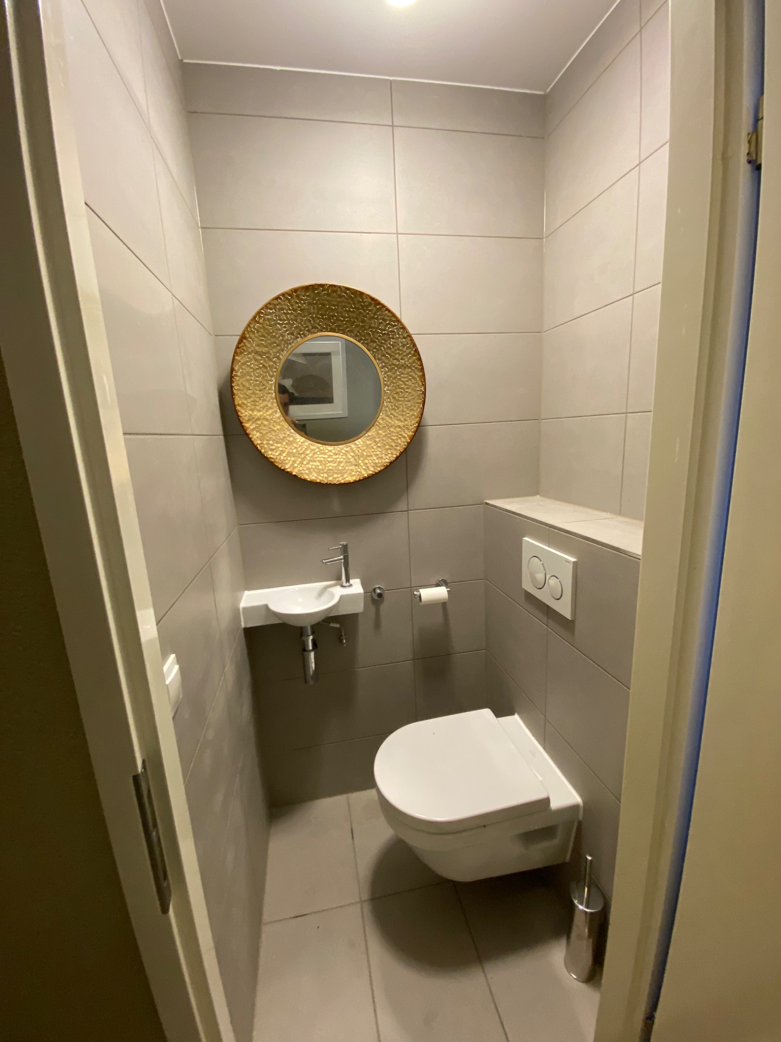 Mała łazienka z szarymi płytkami, okrągłym lustrem w złotej ramie, wiszącą toaletą i małą umywalką.