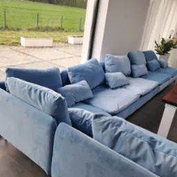 Narożnik modułowy w odcieniach błękitu, z licznymi poduszkami, ustawiony przy oknie z widokiem na zieleń. Nowoczesny design, welurowa tapicerka.