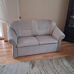 ARISTO-MEBLE KAMILA SZELIGA - Jasnoszara, dwuosobowa sofa tapicerowana w salonie z drewnianą podłogą i dywanem. W tle firanka i ciemna witryna z porcelaną.