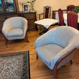 ARISTO-MEBLE KAMILA SZELIGA - Dwa kremowe fotele klubowe z drewnianymi ramami na drewnianej podłodze z dywanem. W tle stół z krzesłami i komoda.