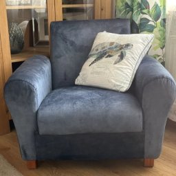 ARISTO-MEBLE KAMILA SZELIGA - Niebieski fotel tapicerowany z poduszką w motywie żółwia, stojący na drewnianej podłodze, z widocznym fragmentem szafki i zasłony w tle.