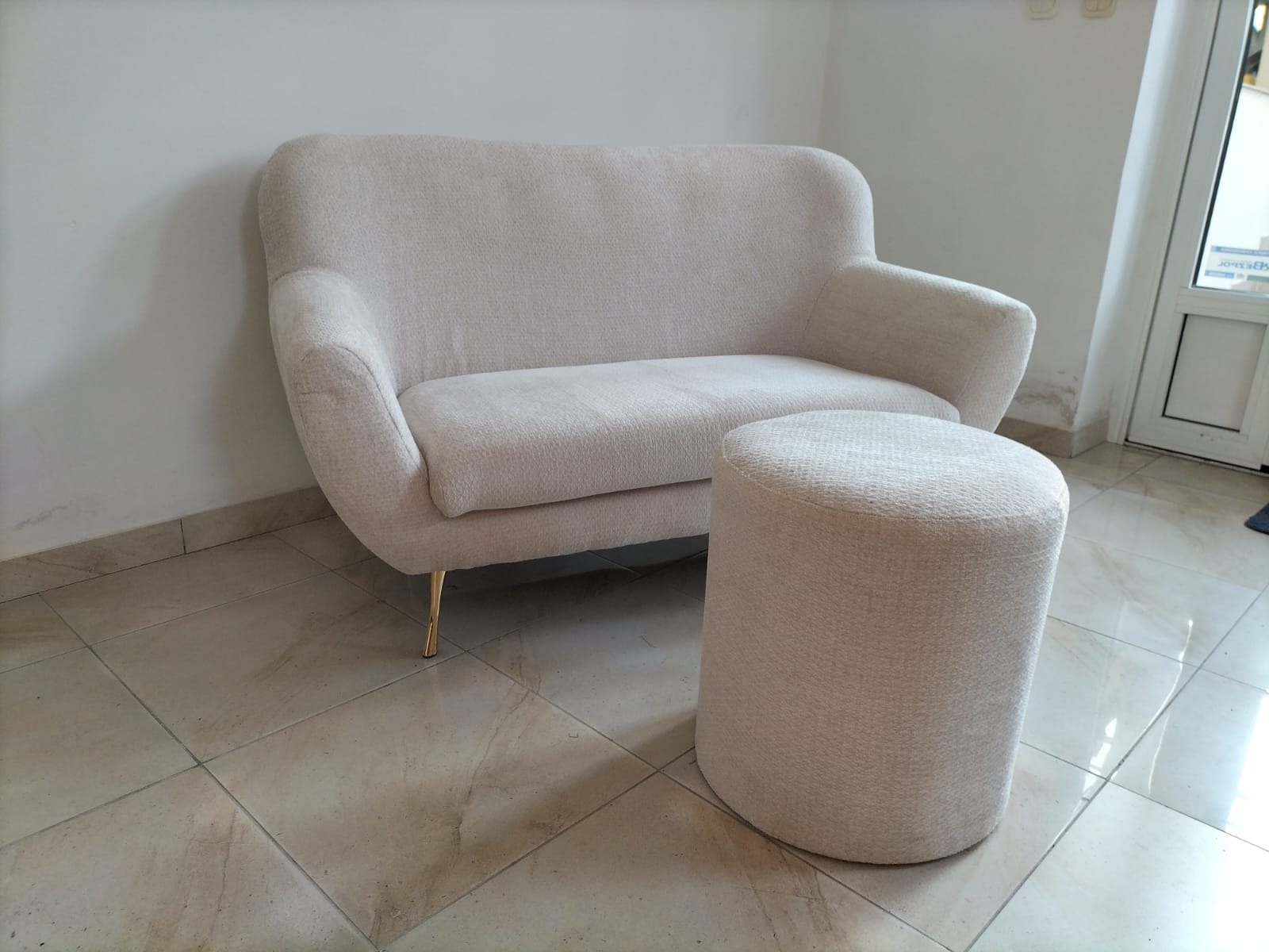 Jasna, dwuosobowa sofa tapicerowana z podnóżkiem. Sofa na złotych nóżkach, w tle biała ściana i drzwi. Nowoczesny design, neutralna kolorystyka.