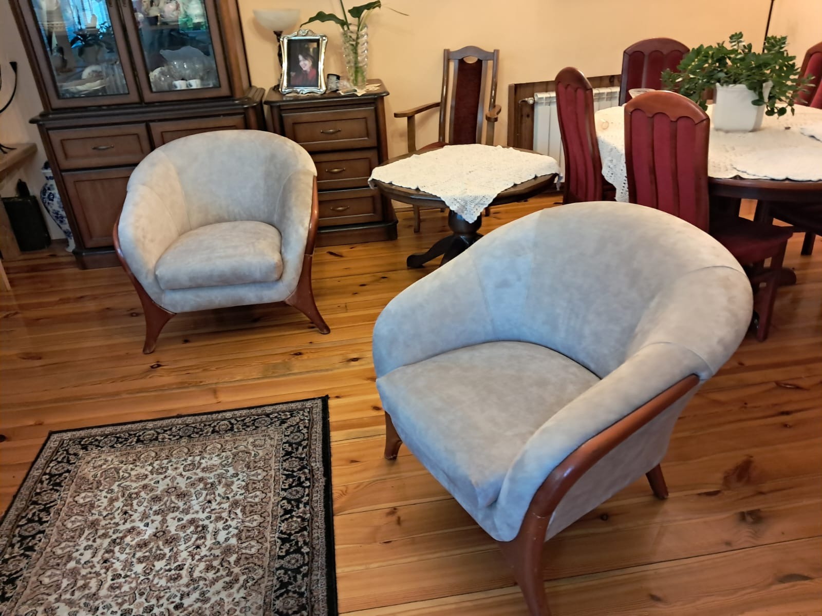 Dwa kremowe fotele klubowe z drewnianymi ramami na drewnianej podłodze z dywanem. W tle stół z krzesłami i komoda.