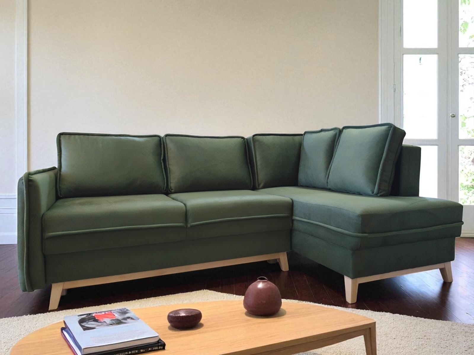 Stylowy narożnik w odcieniach zieleni z poduszkami, na drewnianych nóżkach, ustawiony w jasnym salonie. Na stoliku książki i dekoracje.