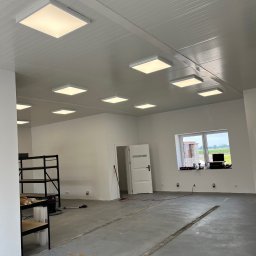 Leszek Lorek ELEKTRO-LO - Wnętrze nowo wykończonego, jasnego pomieszczenia z zamontowanymi kwadratowymi panelami LED w suficie, widoczne regały metalowe, okno z widokiem na zewnątrz i betonowa posadzka z dylatacjami.