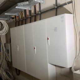 Leszek Lorek ELEKTRO-LO - Rozdzielnia elektryczna z kilkoma białymi szafkami, widoczne kable w korytkach pod sufitem, instalacja w surowym stanie.