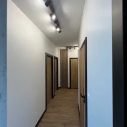 Nowoczesny korytarz z drewnianą podłogą, białymi ścianami, czarnymi futrynami i listwami przypodłogowymi. Na suficie szyna z reflektorami.