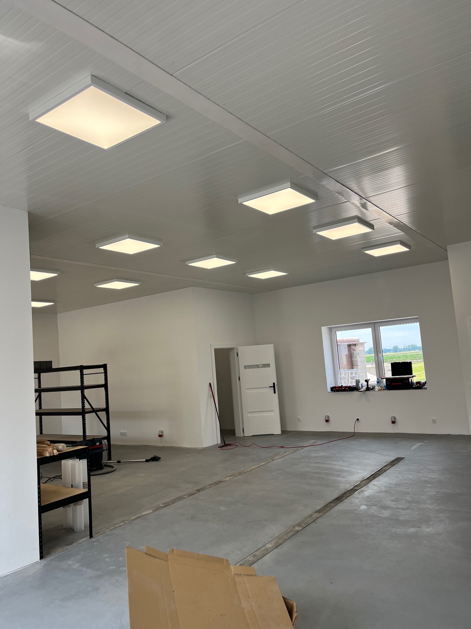 Wnętrze nowo wykończonego, jasnego pomieszczenia z zamontowanymi kwadratowymi panelami LED w suficie, widoczne regały metalowe, okno z widokiem na zewnątrz i betonowa posadzka z dylatacjami.