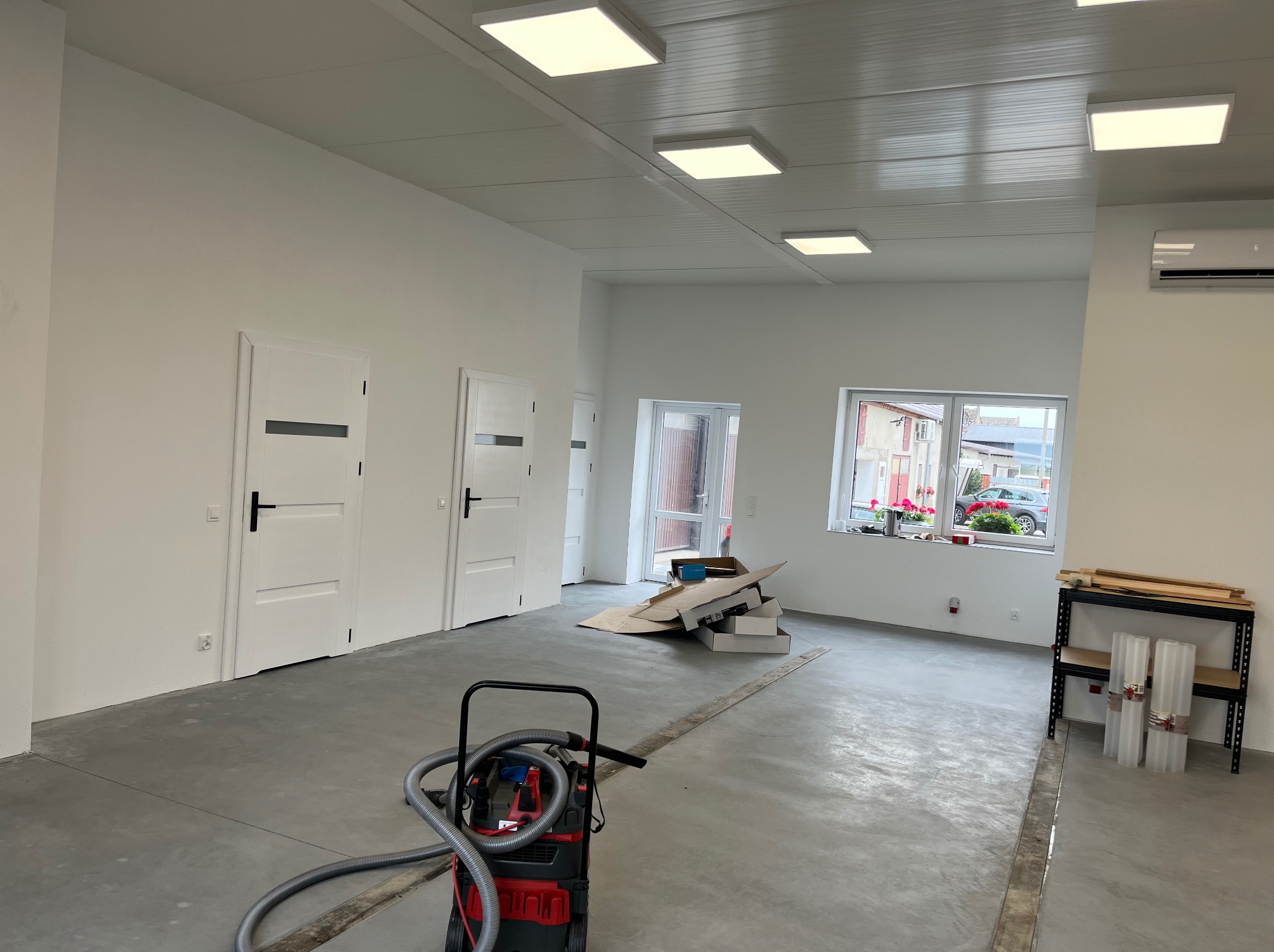 Wnętrze w trakcie wykańczania: surowy beton na podłodze, białe ściany z trzema drzwiami z czarnymi klamkami, sufit z panelami i kwadratowymi lampami, odkurzacz przemysłowy na pierwszym planie, okno...
