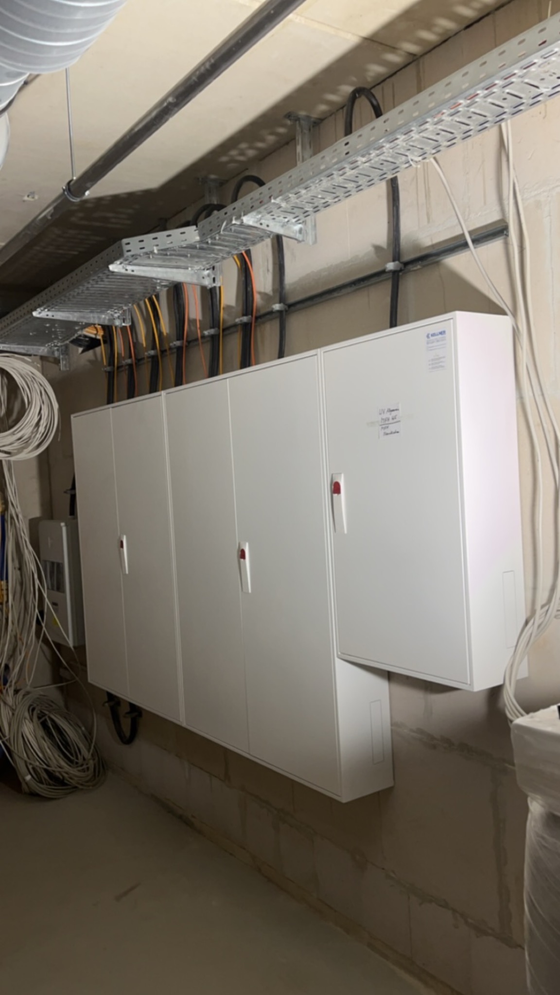 Rozdzielnia elektryczna z kilkoma białymi szafkami, widoczne kable w korytkach pod sufitem, instalacja w surowym stanie.