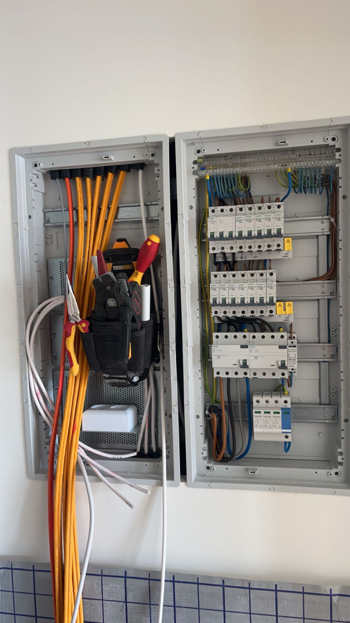 Otwarta skrzynka elektryczna z przewodami, bezpiecznikami i narzędziami w kaburze po lewej stronie, widok z bliska na instalację elektryczną w trakcie prac.