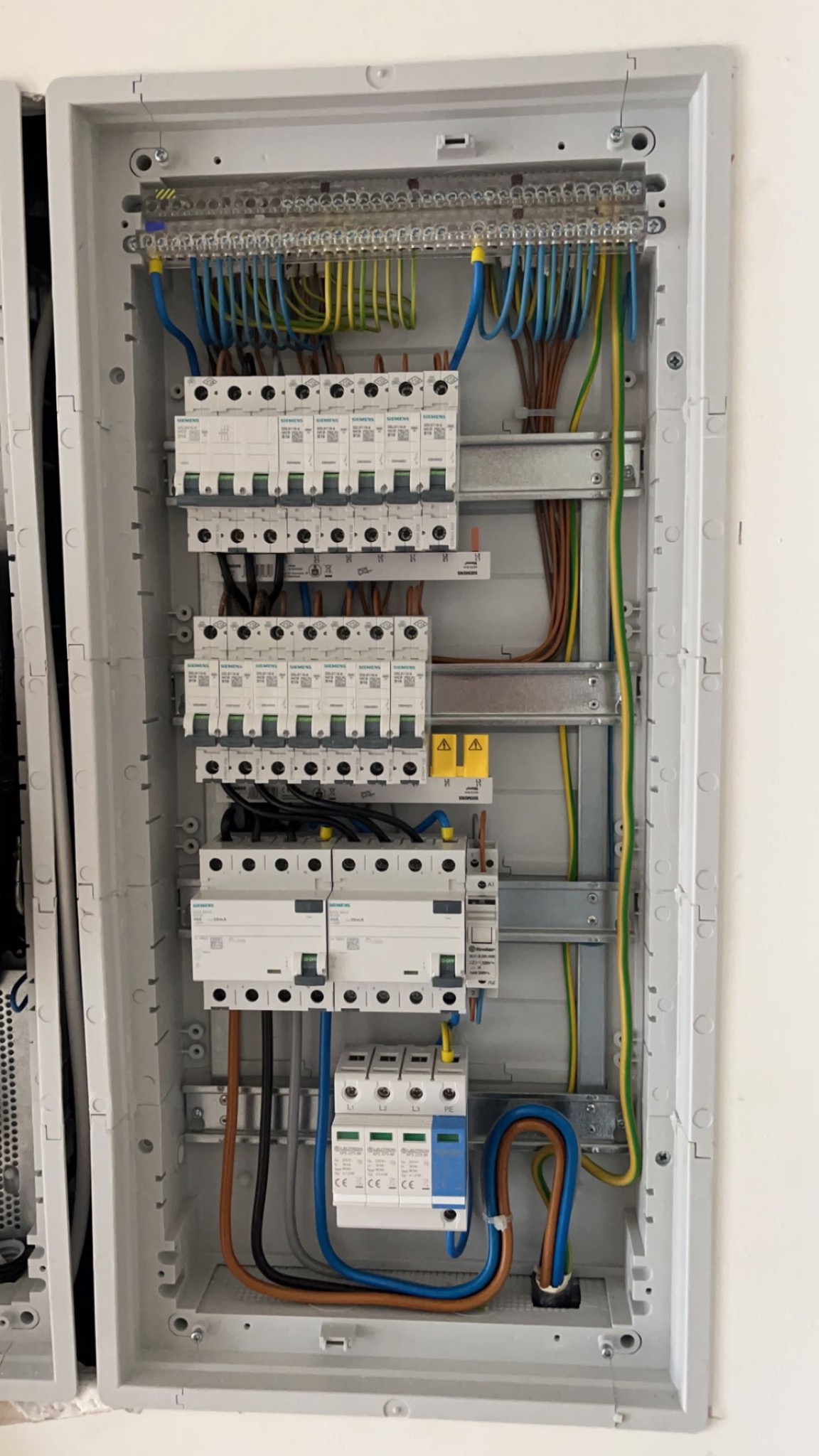 Otwarta skrzynka elektryczna z bezpiecznikami, przewodami w kolorach niebieskim, brązowym, czarnym i żółto-zielonym, zamontowana na białej ścianie.