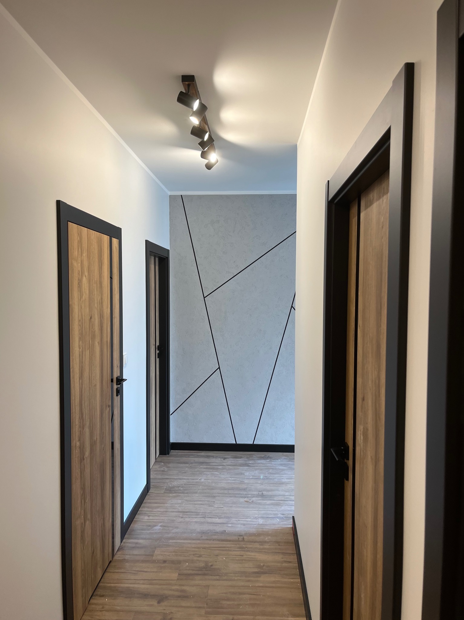 Nowoczesny korytarz z drewnianą podłogą i oświetleniem punktowym. Na ścianie dekoracyjna tapeta z geometrycznym wzorem. Widoczne drzwi z czarnymi ramami i drewnianym wypełnieniem.