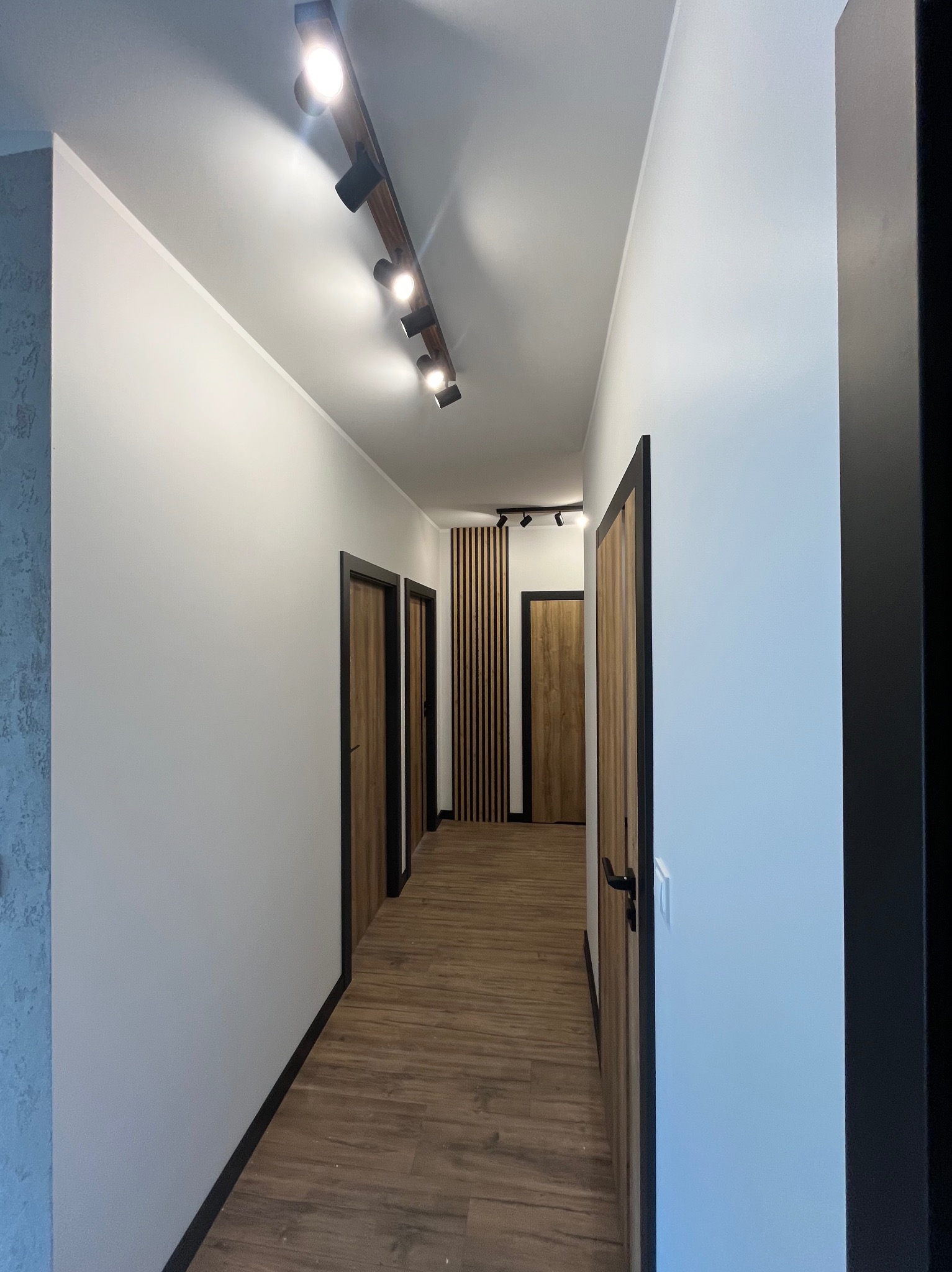Nowoczesny korytarz z drewnianą podłogą, białymi ścianami, czarnymi futrynami i listwami przypodłogowymi. Na suficie szyna z reflektorami.