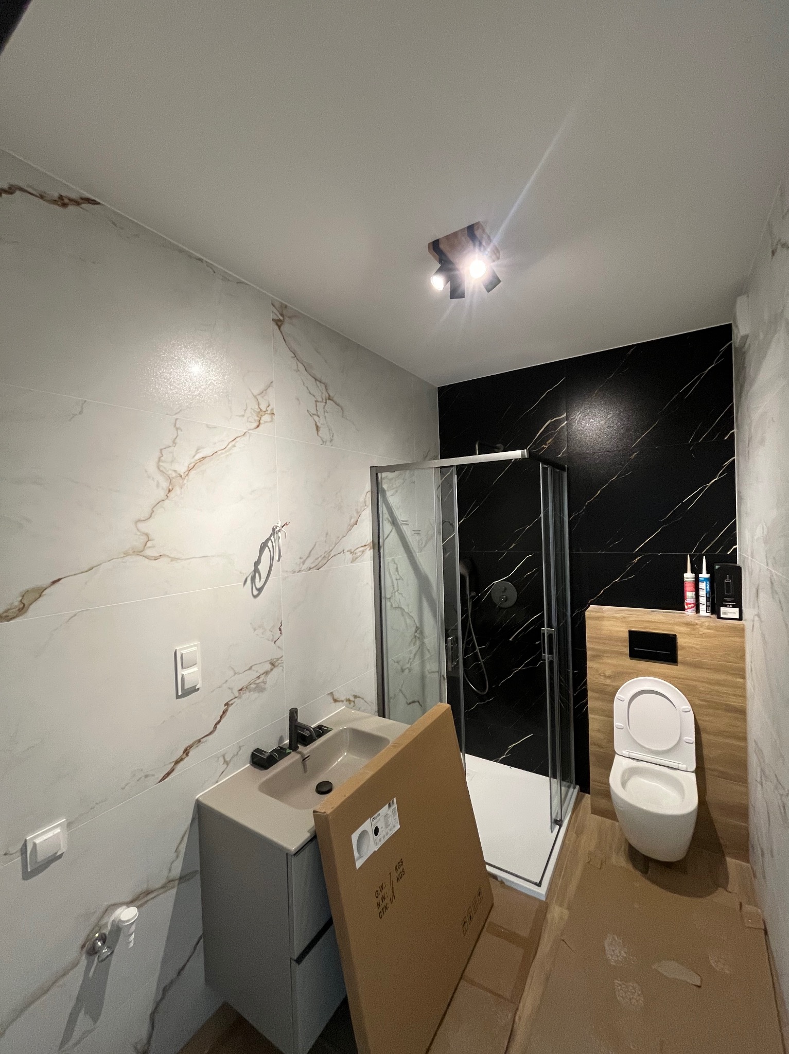Wykończona łazienka z kabiną prysznicową, umywalką i toaletą. Białe i czarne marmurowe płytki. Na podłodze kartonowe zabezpieczenie.