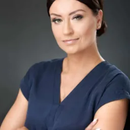 Portret kobiety w granatowej bluzce z założonymi rękami, zegarkiem na nadgarstku i delikatnym uśmiechem na tle szarego studia.