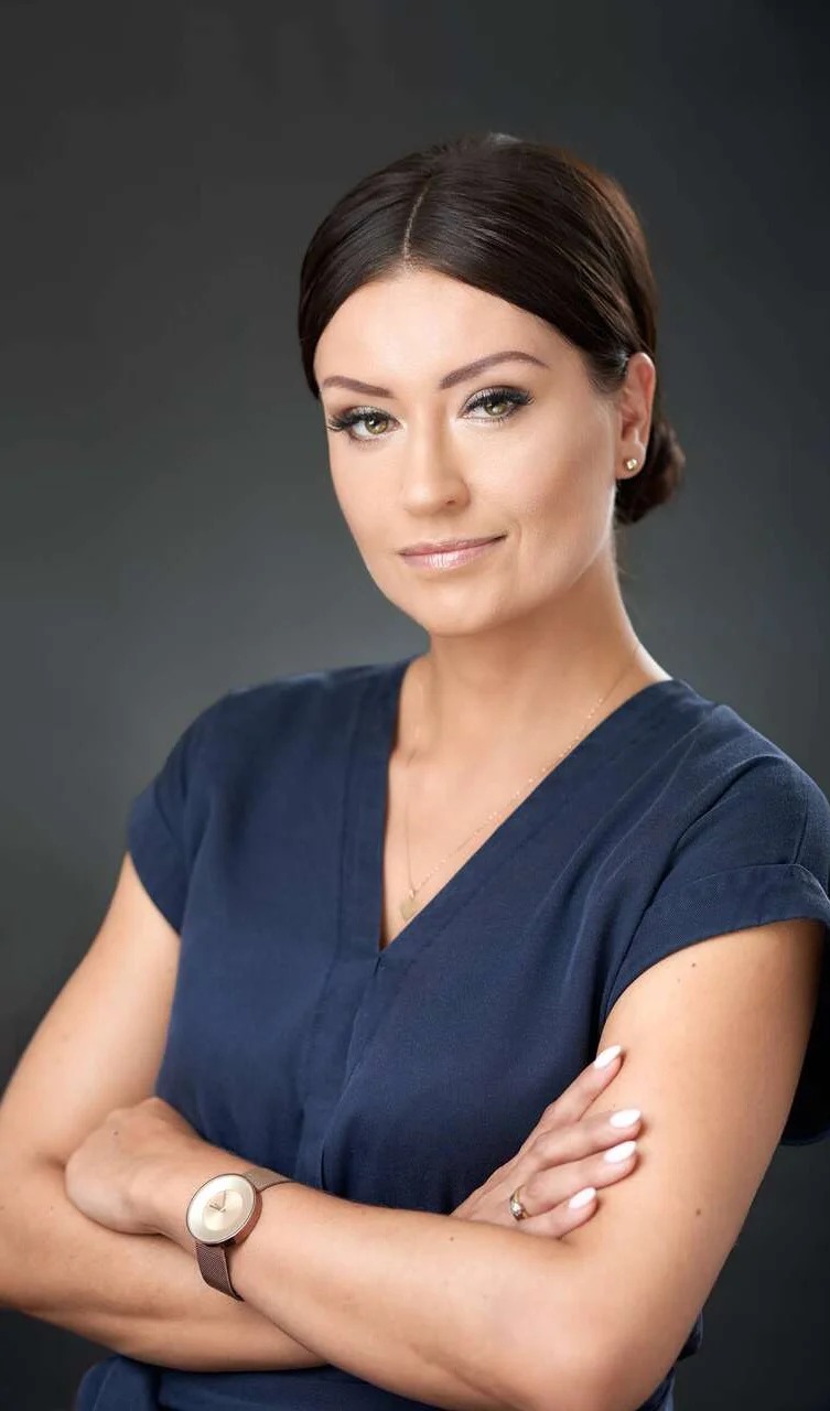 Portret kobiety w granatowej bluzce z założonymi rękami, zegarkiem na nadgarstku i delikatnym uśmiechem na tle szarego studia.