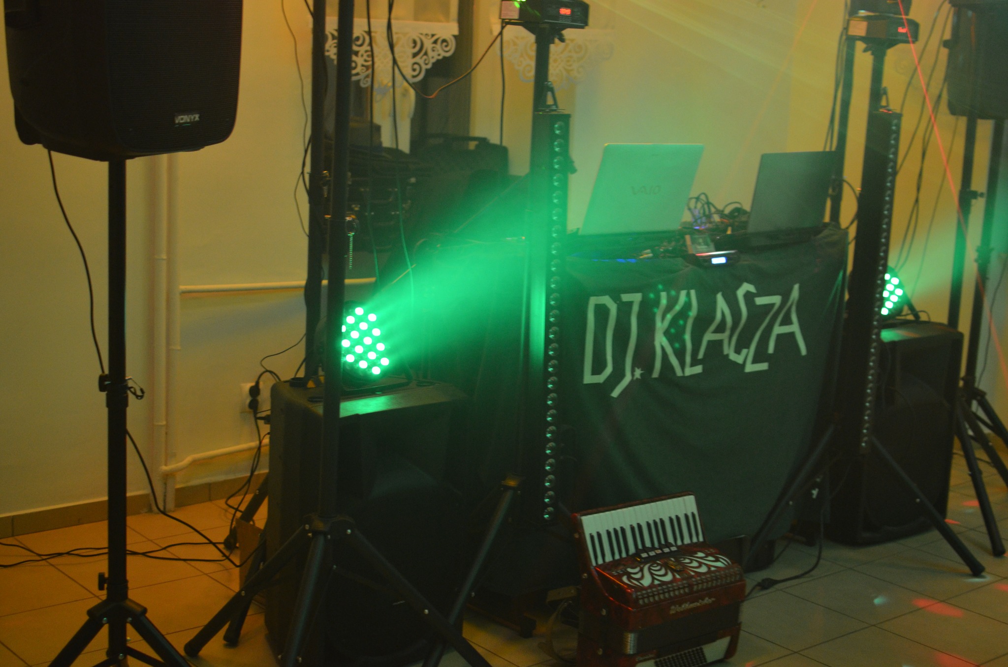 Stanowisko DJ'a z oświetleniem, laptopem i akordeonem na tle ściany. Zielone światło reflektora tworzy atmosferę imprezy.