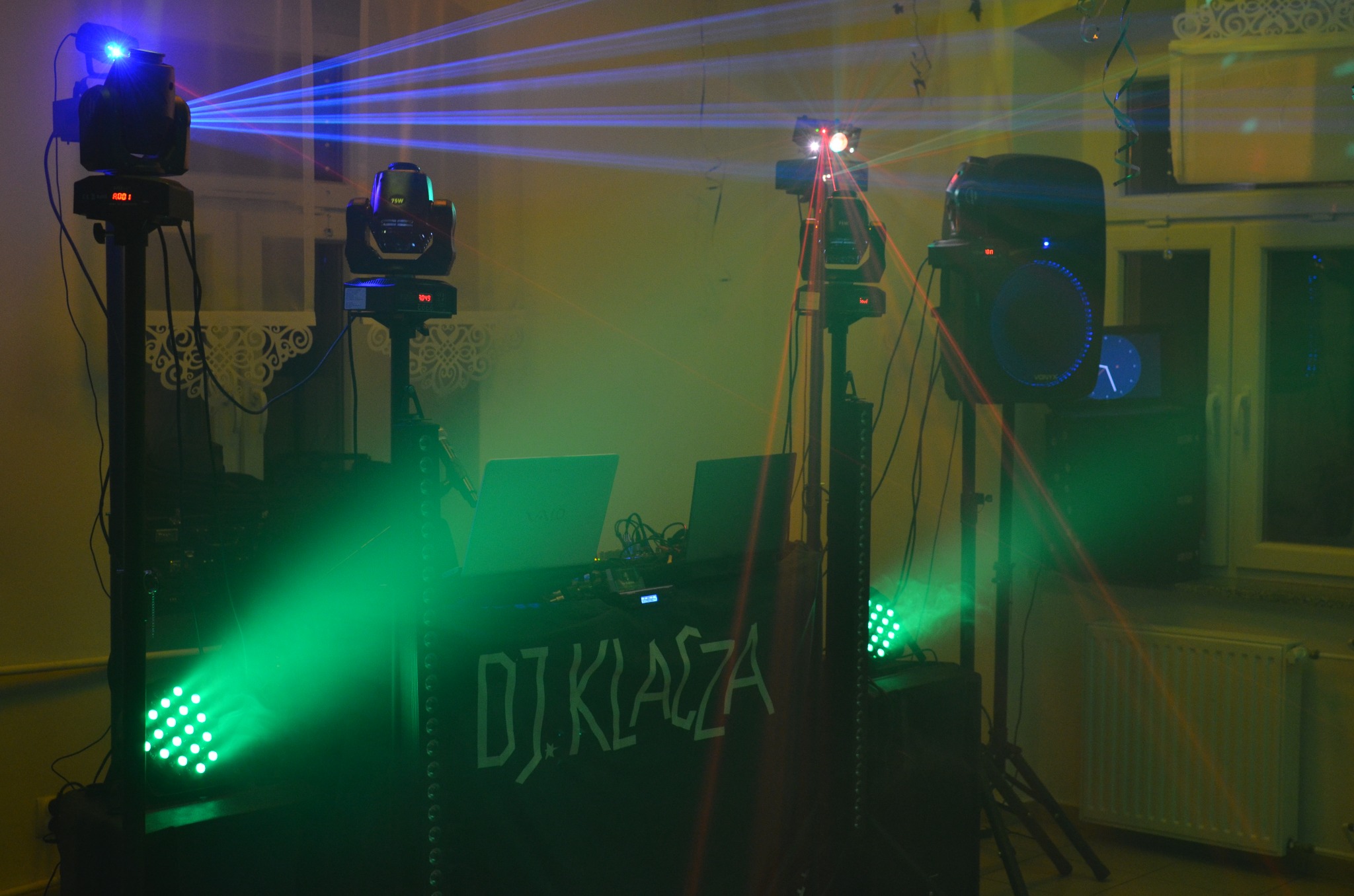 Oświetlenie dyskotekowe z laserami i reflektorami na statywach, z napisem 'DJ KLACZA' na froncie, tworzące imprezową atmosferę w pomieszczeniu.