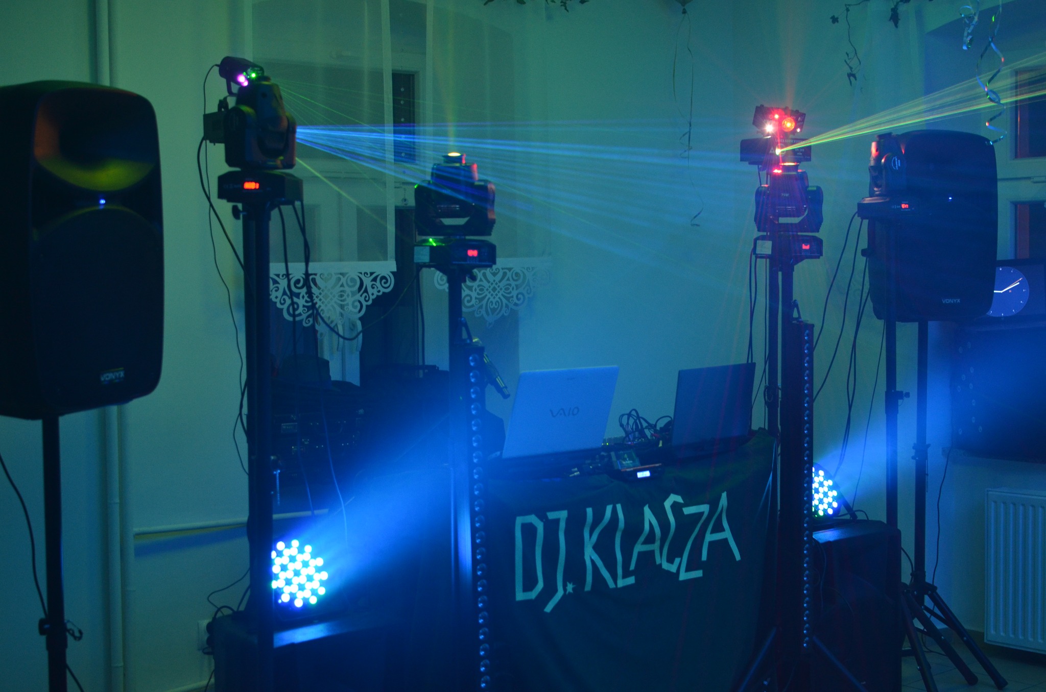 Scena DJ-a z oświetleniem dyskotekowym, laptopem VAIO i głośnikami Vonyx. Kolorowe lasery tworzą atmosferę imprezy. Widoczne kable i dekoracje.