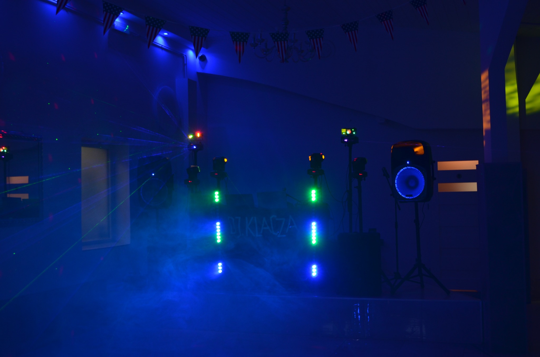 Oświetlenie sceniczne z laserami i kolumną głośnikową w niebieskiej poświacie, dekoracje z flag USA w tle. DJ Klacza na scenie.